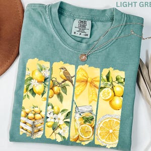 May include: Light green t-shirt with a lemon-themed graphic. The design features yellow lemons, a bird, a yellow bow, and a mason jar with lemons. The shirt has a Comfort Colors tag and a silver necklace.