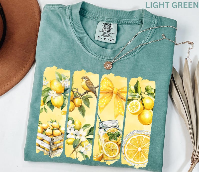 May include: Light green t-shirt with a lemon-themed graphic. The design features yellow lemons, a bird, a yellow bow, and a mason jar with lemons. The shirt has a Comfort Colors tag and a silver necklace.