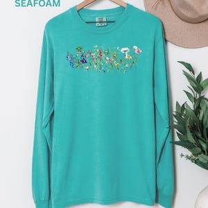 May include: Seafoam green long-sleeve shirt with a floral design across the chest. The design features a variety of colorful wildflowers. The word "SEAFOAM" is printed in the upper left corner of the image.