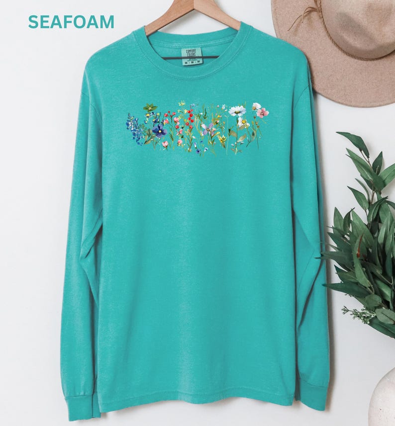May include: Seafoam green long-sleeve shirt with a floral design across the chest. The design features a variety of colorful wildflowers. The word "SEAFOAM" is printed in the upper left corner of the image.