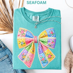 May include: Seafoam green t-shirt with a colourful patchwork bow design. The bow features floral, polka dot, and gingham patterns in pink, yellow, blue, and purple. A silver necklace with a pendant is on the shirt.