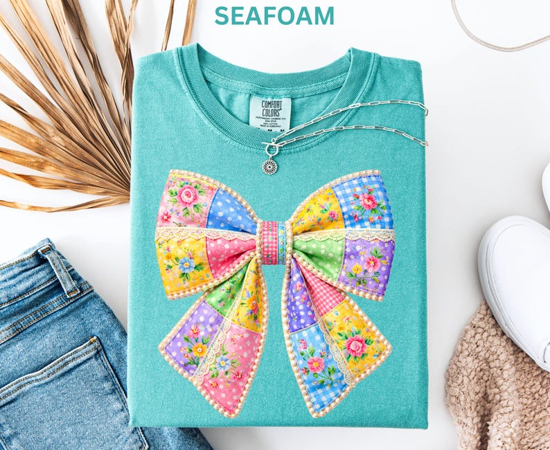 May include: Seafoam green t-shirt with a colourful patchwork bow design. The bow features floral, polka dot, and gingham patterns in pink, yellow, blue, and purple. A silver necklace with a pendant is on the shirt.