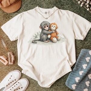 Puede incluir: Camiseta de color crema con una ilustraci&oacute;n en acuarela de dos monos abraz&aacute;ndose. Un mono es gris, el otro es naranja. La camiseta tiene cuello redondo y mangas cortas. El dise&ntilde;o est&aacute; centrado en la parte delantera.