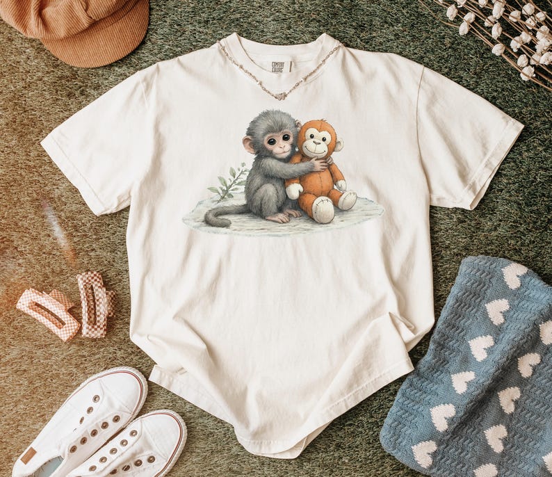 Puede incluir: Camiseta de color crema con una ilustraci&oacute;n en acuarela de dos monos abraz&aacute;ndose. Un mono es gris, el otro es naranja. La camiseta tiene cuello redondo y mangas cortas. El dise&ntilde;o est&aacute; centrado en la parte delantera.