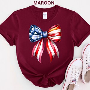 May include: Maroon t-shirt featuring a watercolor bow design in red, white, and blue, resembling the American flag. The bow has a blue section with white stars and red and white stripes. White sneakers are placed near the shirt.