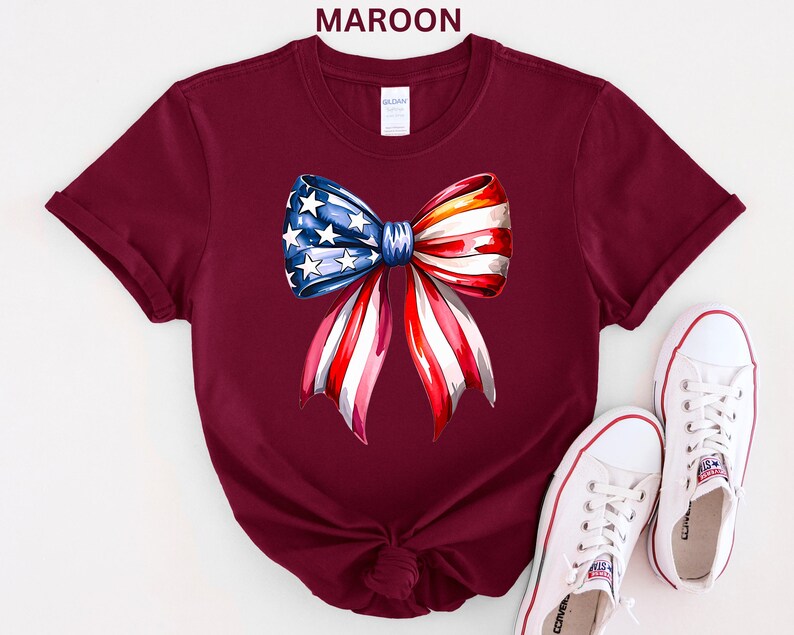 May include: Maroon t-shirt featuring a watercolor bow design in red, white, and blue, resembling the American flag. The bow has a blue section with white stars and red and white stripes. White sneakers are placed near the shirt.