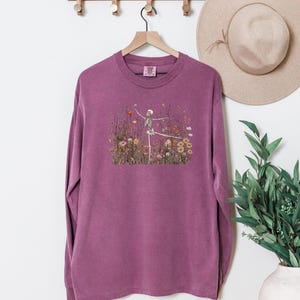 Puede incluir: Una camiseta de manga larga con cuello redondo en color ciruela apagado. El centro del pecho presenta un estampado gr&aacute;fico de un esqueleto humano en pose de baile, rodeado por una ilustraci&oacute;n detallada de flores silvestres y mariposas.