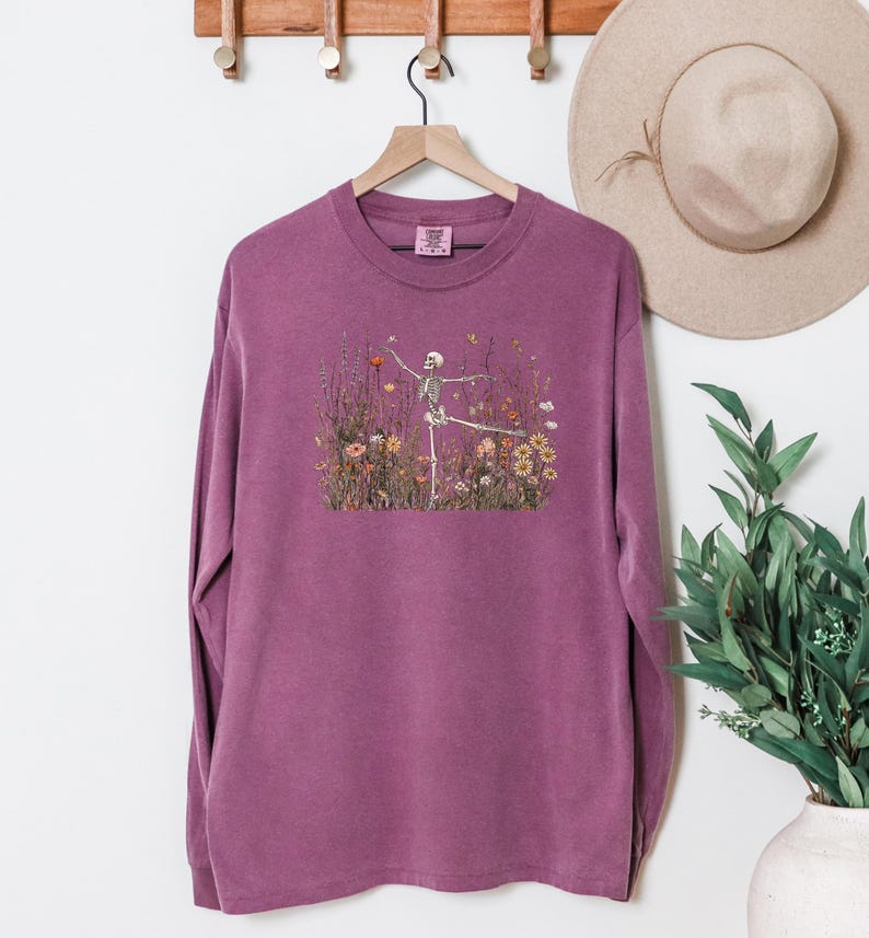 Puede incluir: Una camiseta de manga larga con cuello redondo en color ciruela apagado. El centro del pecho presenta un estampado gr&aacute;fico de un esqueleto humano en pose de baile, rodeado por una ilustraci&oacute;n detallada de flores silvestres y mariposas.