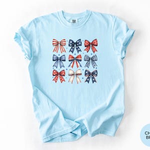 May include: A light blue t-shirt featuring a central graphic print of nine decorative bows arranged in a three-by-three grid. The bows display various patterns including red and white stripes, blue with white stars, and red or blue polka dots.
