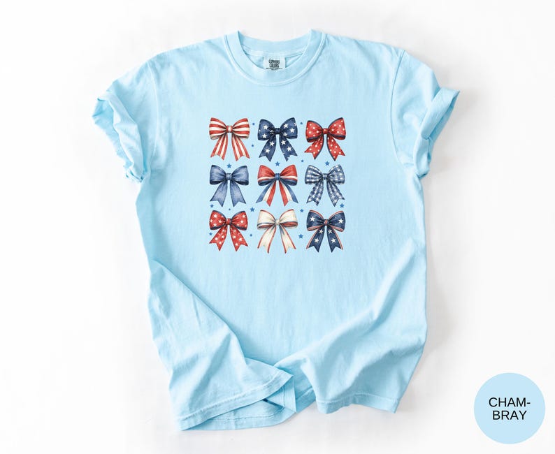 May include: A light blue t-shirt featuring a central graphic print of nine decorative bows arranged in a three-by-three grid. The bows display various patterns including red and white stripes, blue with white stars, and red or blue polka dots.