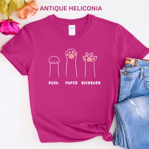 Puede incluir: Una camiseta magenta con un dise&ntilde;o l&uacute;dico que presenta ilustraciones en forma de pata que representan piedra, papel y tijeras. El texto "ROCK PAPER SCISSORS" est&aacute; impreso debajo. La camiseta se combina con unos vaqueros azules.