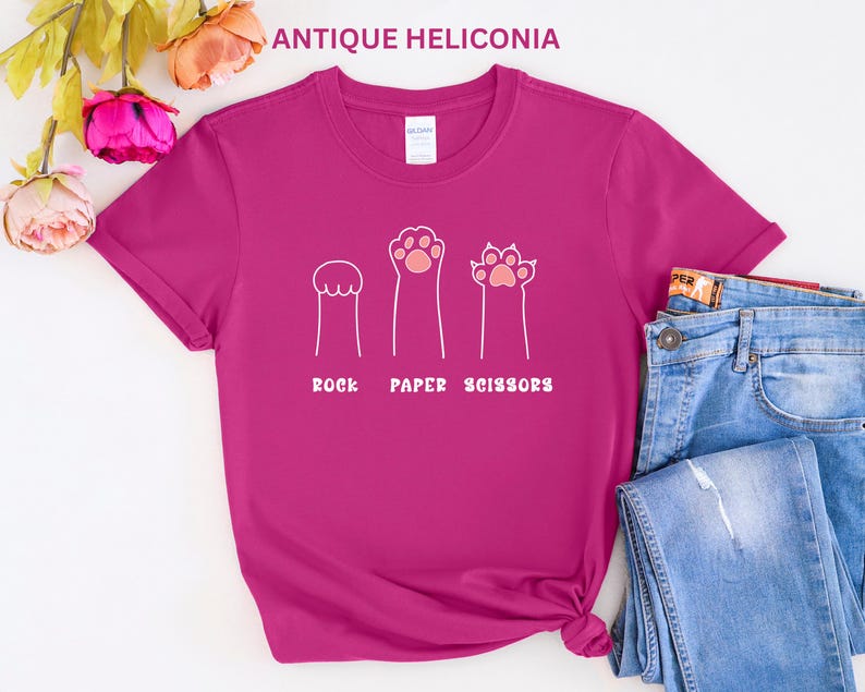 Puede incluir: Una camiseta magenta con un dise&ntilde;o l&uacute;dico que presenta ilustraciones en forma de pata que representan piedra, papel y tijeras. El texto "ROCK PAPER SCISSORS" est&aacute; impreso debajo. La camiseta se combina con unos vaqueros azules.