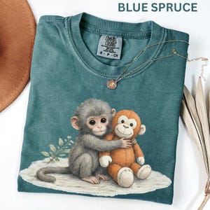 Puede incluir: Camiseta Comfort Colors azul abeto con un gr&aacute;fico de un mono gris abrazando a un mono de peluche naranja. La camiseta tiene cuello redondo y mangas cortas. Un collar dorado con un peque&ntilde;o colgante est&aacute; sobre la camiseta.