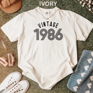 May include: Ivory-colored t-shirt with the text "VINTAGE 1986" in black. The shirt has a classic crew neck and short sleeves. The graphic features a retro design. The shirt is laid flat on a textured surface.