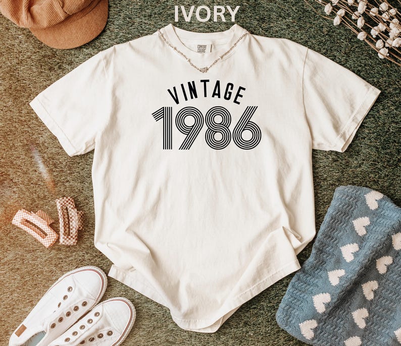 May include: Ivory-colored t-shirt with the text "VINTAGE 1986" in black. The shirt has a classic crew neck and short sleeves. The graphic features a retro design. The shirt is laid flat on a textured surface.