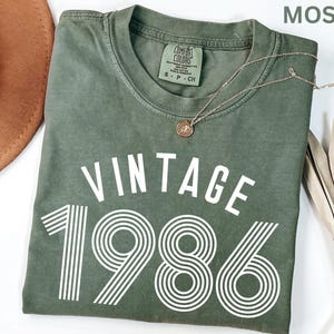 May include: A moss green t-shirt with the word "VINTAGE" above the number "1986" in white. A silver necklace with a pendant rests on the shirt. The shirt has a crew neck and a small tag on the upper left.