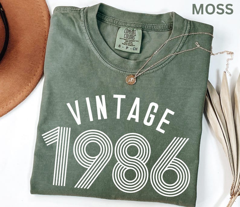 May include: A moss green t-shirt with the word "VINTAGE" above the number "1986" in white. A silver necklace with a pendant rests on the shirt. The shirt has a crew neck and a small tag on the upper left.