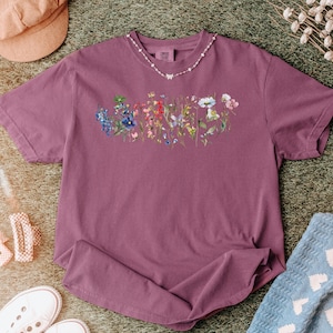May include: A mauve-colored t-shirt featuring a floral design across the chest. The design showcases a variety of colorful wildflowers. A delicate necklace with a butterfly pendant adorns the neckline. The shirt is laid out on a green surface.