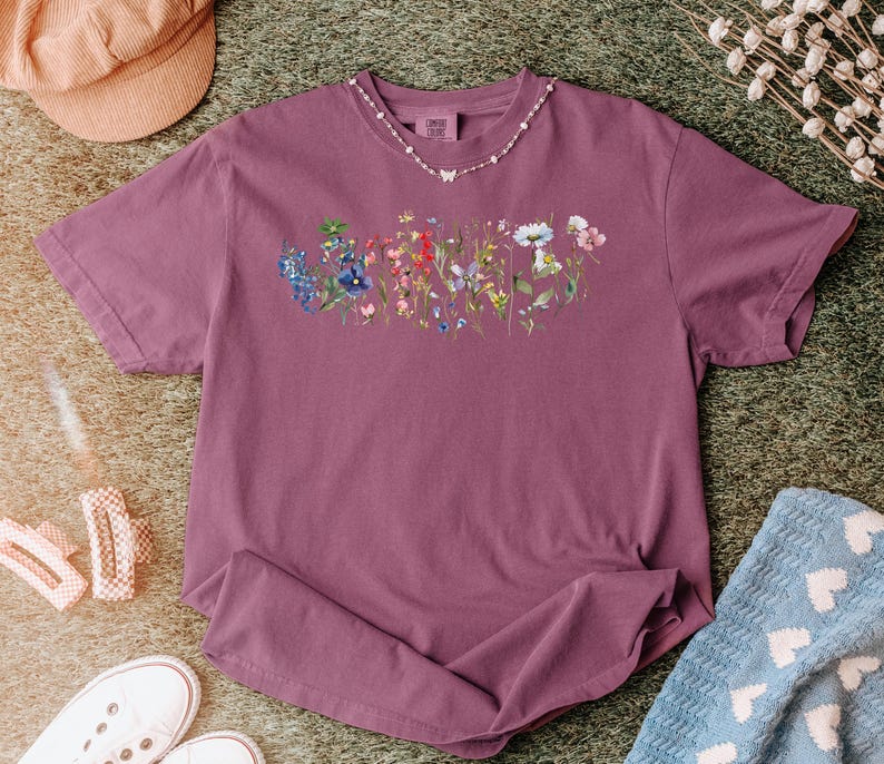 May include: A mauve-colored t-shirt featuring a floral design across the chest. The design showcases a variety of colorful wildflowers. A delicate necklace with a butterfly pendant adorns the neckline. The shirt is laid out on a green surface.