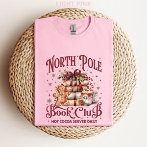 A pink t-shirt with a graphic design featuring a gingerbread man, books, and a Christmas tree, along with text that says "North Pole Book Club" and "Hot Cocoa Served Daily".