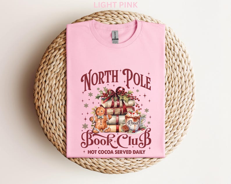 A pink t-shirt with a graphic design featuring a gingerbread man, books, and a Christmas tree, along with text that says "North Pole Book Club" and "Hot Cocoa Served Daily".