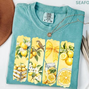 May include: Seafoam green Comfort Colors t-shirt with a lemon-themed graphic. The graphic features lemons, a bird, a yellow bow, and a glass of lemonade. The shirt has a crew neck and short sleeves. A silver necklace with a pendant is also visible.