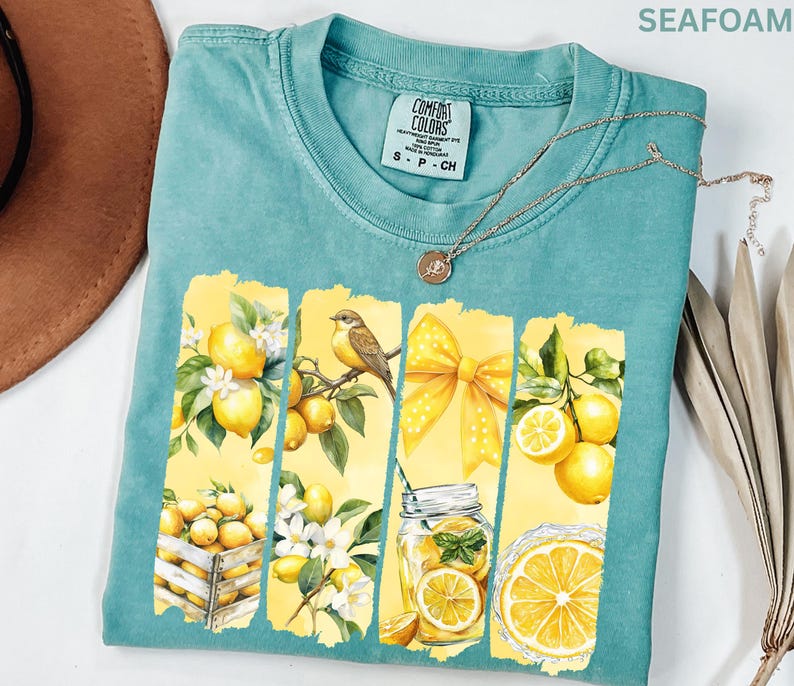 May include: Seafoam green Comfort Colors t-shirt with a lemon-themed graphic. The graphic features lemons, a bird, a yellow bow, and a glass of lemonade. The shirt has a crew neck and short sleeves. A silver necklace with a pendant is also visible.