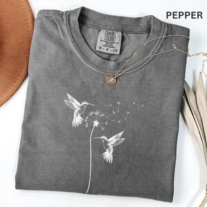 a gray t-shirt with a graphic design of two hummingbirds, along with a necklace and a hat.