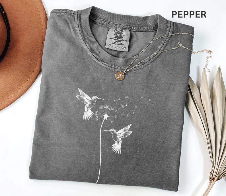 a gray t-shirt with a graphic design of two hummingbirds, along with a necklace and a hat.