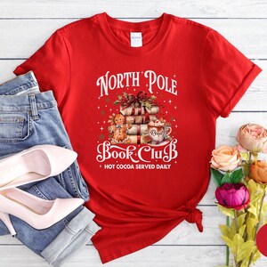 May include: A red short-sleeved t-shirt featuring a graphic of a gingerbread person, a stack of books, and a mug of hot cocoa. The text reads North Pole Book Club and Hot Cocoa Served Daily. The shirt is paired with blue denim jeans and heels.