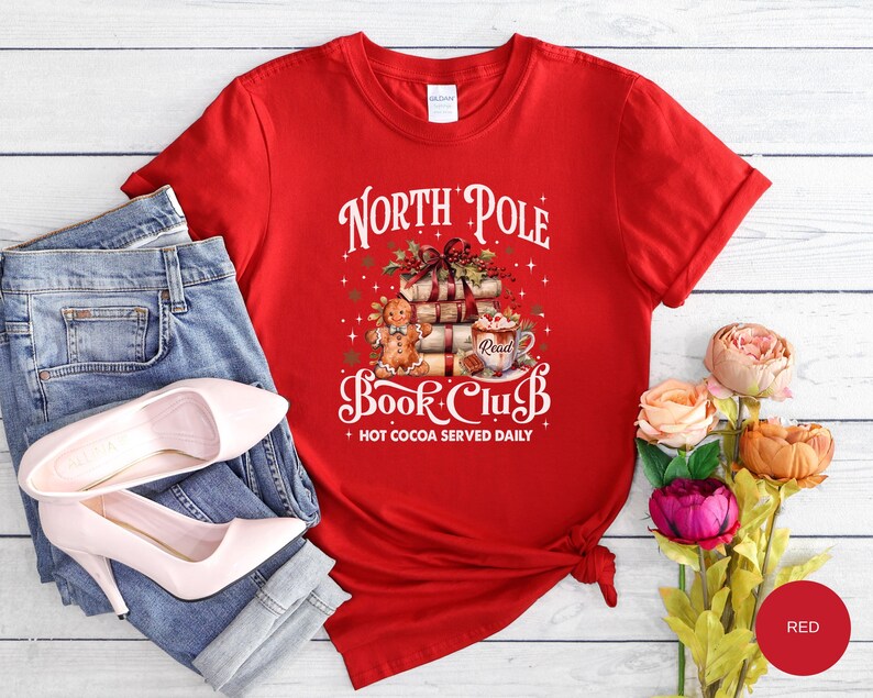 May include: A red short-sleeved t-shirt featuring a graphic of a gingerbread person, a stack of books, and a mug of hot cocoa. The text reads North Pole Book Club and Hot Cocoa Served Daily. The shirt is paired with blue denim jeans and heels.