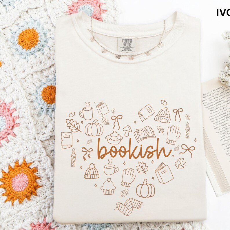 A white t-shirt with a heart-shaped design featuring the word "bookish" and various fall-themed illustrations, such as pumpkins, leaves, and mugs, is displayed on a white surface.