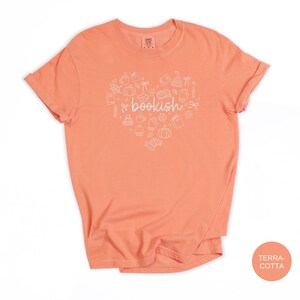May include: A terracotta-colored short-sleeve t-shirt featuring a white graphic print in the shape of a heart. The heart contains various line-art icons including books, pumpkins, mugs, and leaves, with the word bookish centered in the middle.