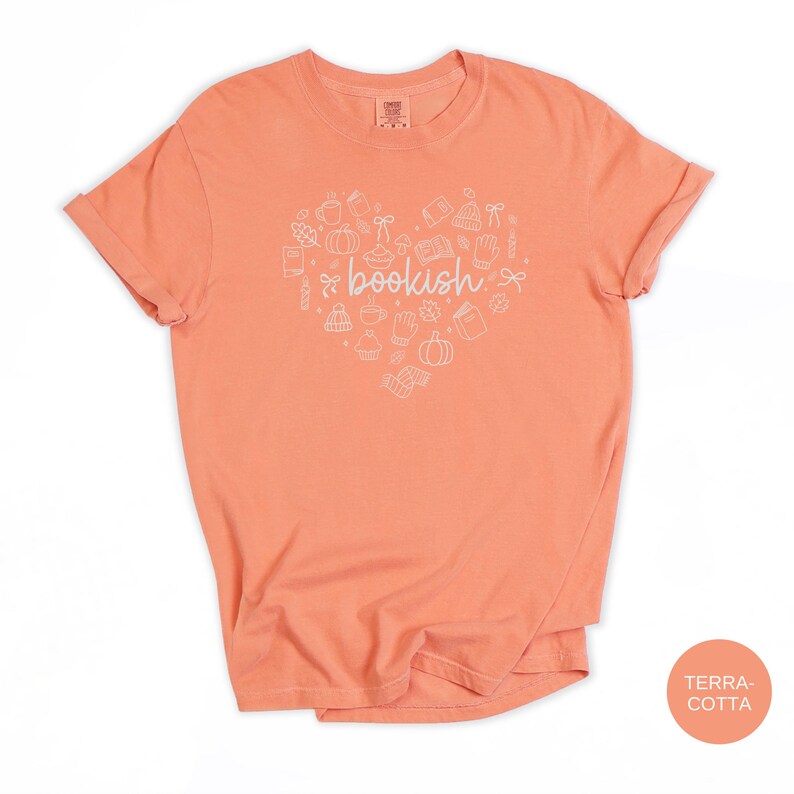 May include: A terracotta-colored short-sleeve t-shirt featuring a white graphic print in the shape of a heart. The heart contains various line-art icons including books, pumpkins, mugs, and leaves, with the word bookish centered in the middle.