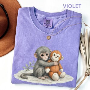 Puede incluir: Camiseta violeta Comfort Colors con un gr&aacute;fico de un mono gris abrazando a un mono de peluche naranja. Tambi&eacute;n se ven un collar dorado y un sombrero marr&oacute;n. La camiseta tiene una peque&ntilde;a etiqueta que dice "Comfort Colors".