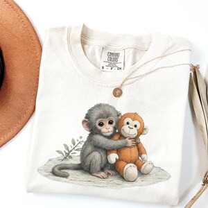 Puede incluir: Camiseta Comfort Colors color crema con una ilustraci&oacute;n en acuarela de dos monos abraz&aacute;ndose. Uno es gris, el otro naranja. Tambi&eacute;n se ven un collar dorado con un colgante y un sombrero marr&oacute;n.