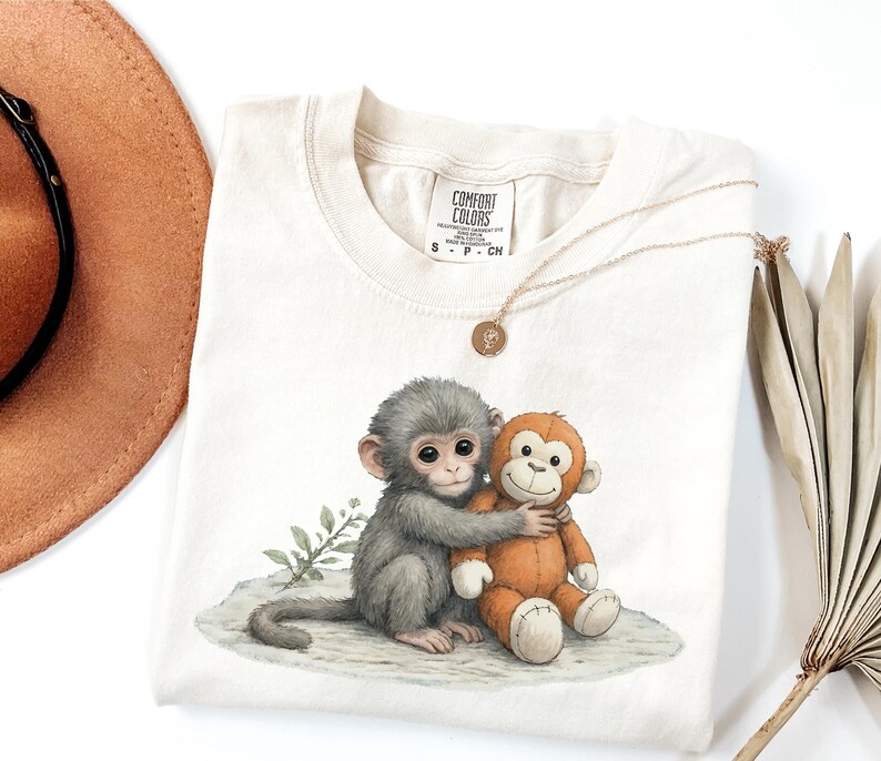 Puede incluir: Camiseta Comfort Colors color crema con una ilustraci&oacute;n en acuarela de dos monos abraz&aacute;ndose. Uno es gris, el otro naranja. Tambi&eacute;n se ven un collar dorado con un colgante y un sombrero marr&oacute;n.