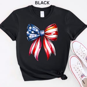 May include: Black t-shirt featuring a watercolor bow design in red, white, and blue, with stars on the blue section. The word "BLACK" is printed at the top. White sneakers with red accents are placed next to the shirt.