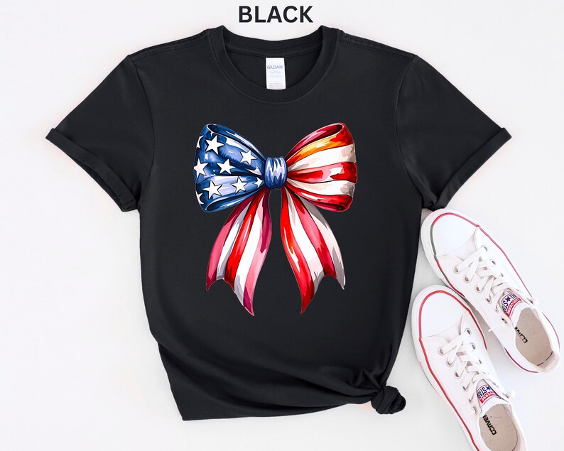 May include: Black t-shirt featuring a watercolor bow design in red, white, and blue, with stars on the blue section. The word "BLACK" is printed at the top. White sneakers with red accents are placed next to the shirt.