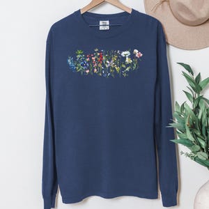 May include: Navy blue long-sleeve shirt with a floral design across the chest. The design features a variety of colorful flowers, including blue, pink, and white blooms. The shirt is hanging on a wooden hanger.