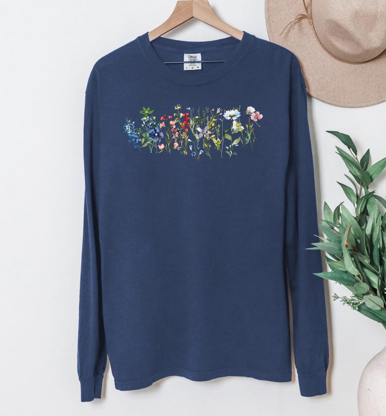 May include: Navy blue long-sleeve shirt with a floral design across the chest. The design features a variety of colorful flowers, including blue, pink, and white blooms. The shirt is hanging on a wooden hanger.