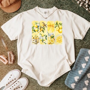 May include: Cream-colored t-shirt featuring a watercolor design of lemons, a bird, and a bow. The design is divided into vertical panels. The shirt is surrounded by accessories, including white sneakers and a brown hat.