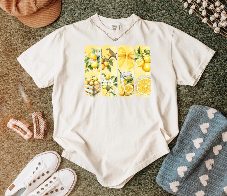 May include: Cream-colored t-shirt featuring a watercolor design of lemons, a bird, and a bow. The design is divided into vertical panels. The shirt is surrounded by accessories, including white sneakers and a brown hat.