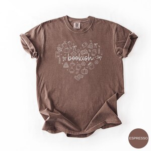May include: A brown short-sleeve t-shirt featuring a white line-art graphic in the shape of a heart. The design includes small illustrations of books, pumpkins, mugs, leaves, and mittens, with the word bookish written in the center.