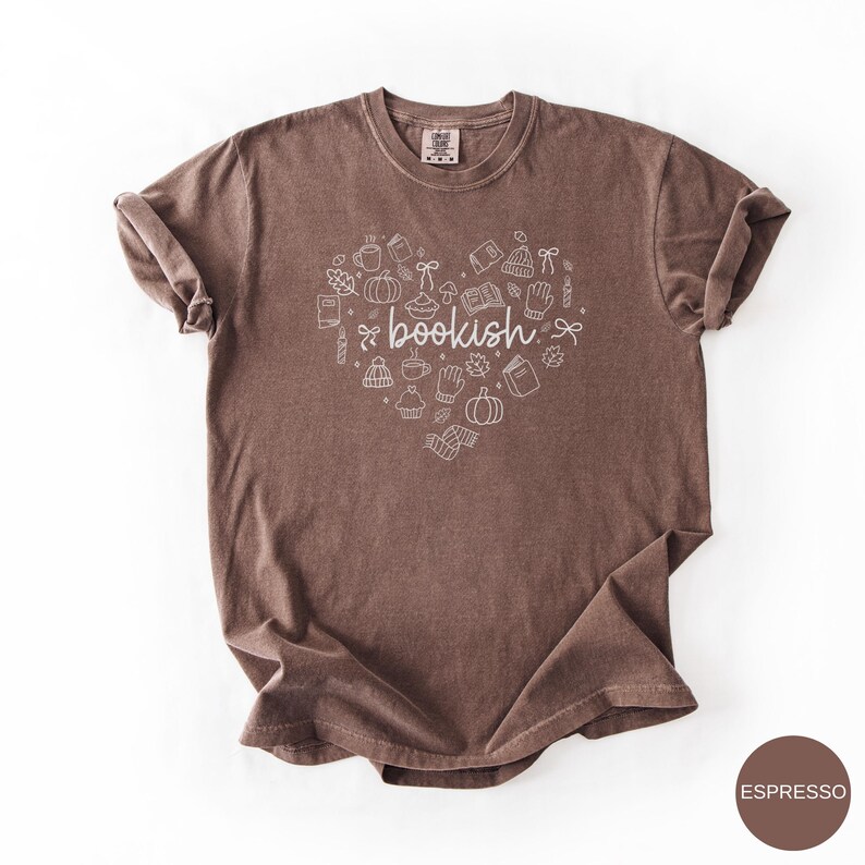 May include: A brown short-sleeve t-shirt featuring a white line-art graphic in the shape of a heart. The design includes small illustrations of books, pumpkins, mugs, leaves, and mittens, with the word bookish written in the center.