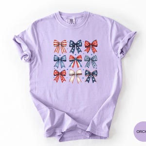 May include: A light purple short-sleeve t-shirt featuring a grid of nine decorative bows in red, white, and blue patterns. The bows include designs with stars, stripes, and polka dots. The shirt has a crew neckline and a relaxed fit.