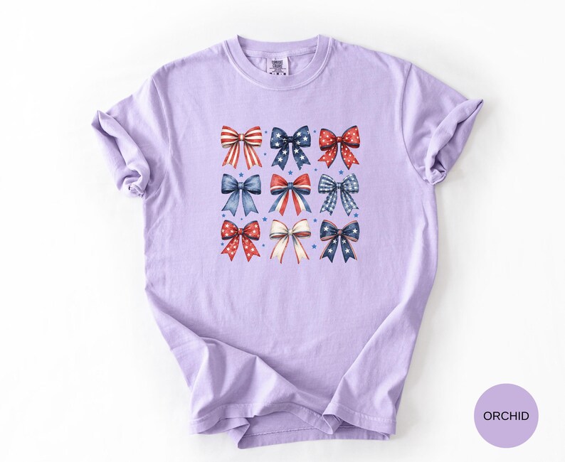 May include: A light purple short-sleeve t-shirt featuring a grid of nine decorative bows in red, white, and blue patterns. The bows include designs with stars, stripes, and polka dots. The shirt has a crew neckline and a relaxed fit.