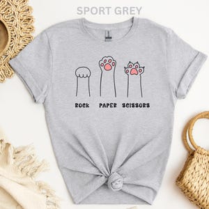 Puede incluir: Camiseta gris deportivo con un divertido dise&ntilde;o de patas de gato que representan piedra, papel y tijeras. El texto "ROCK PAPER SCISSORS" est&aacute; impreso debajo. Una camiseta informal y c&oacute;moda.
