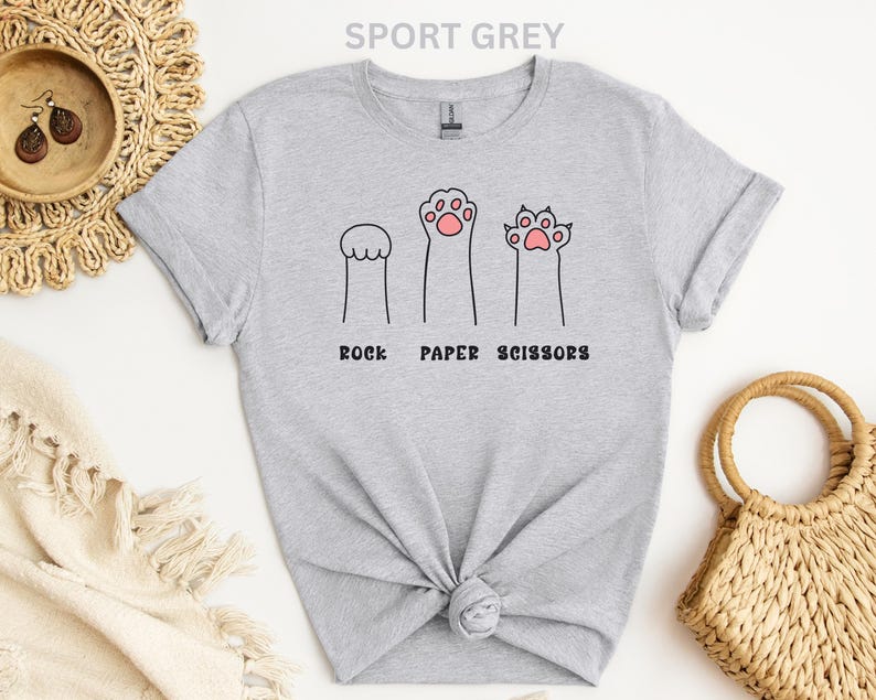 Puede incluir: Camiseta gris deportivo con un divertido dise&ntilde;o de patas de gato que representan piedra, papel y tijeras. El texto "ROCK PAPER SCISSORS" est&aacute; impreso debajo. Una camiseta informal y c&oacute;moda.