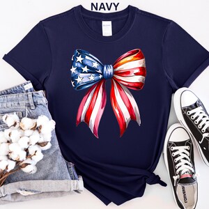 May include: Navy blue t-shirt featuring a watercolor bow design in red, white, and blue, with stars and stripes. The word "NAVY" is printed at the top. The shirt is styled with denim shorts and black sneakers.